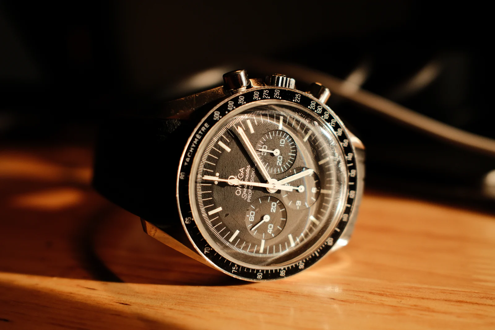 Omega Speedmaster Professional chronograph with black dial and tachymeter bezel, lying on a wooden table in warm side light, black strap fading into shadow.