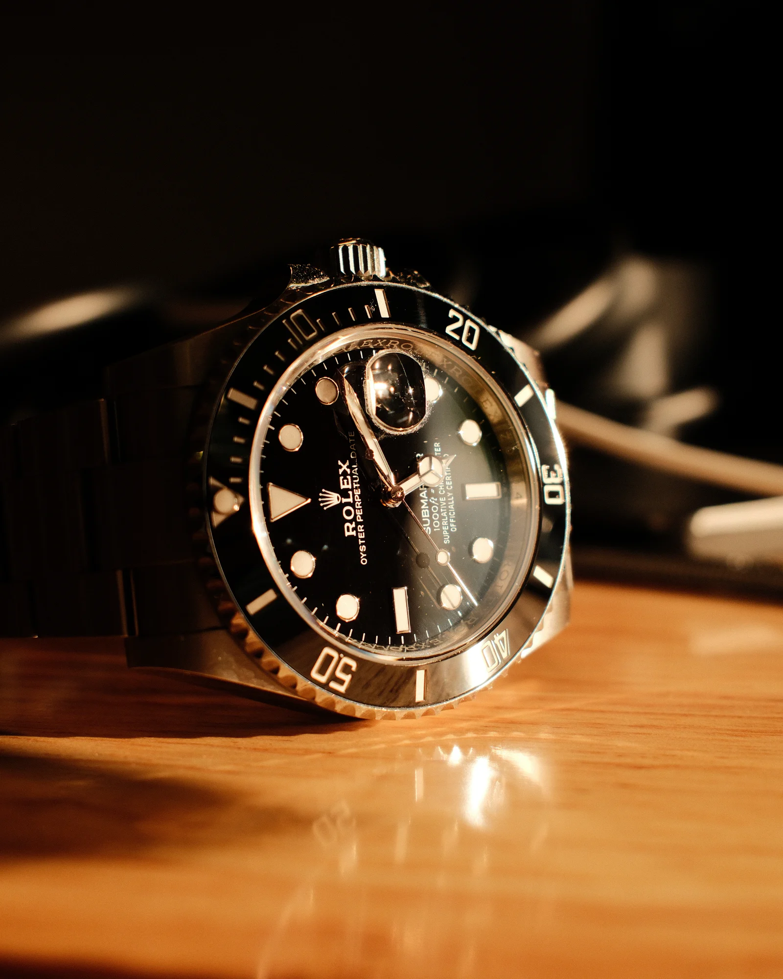 A Rolex Submariner wristwatch with a black dial and black bezel lies on its side on a polished wooden surface, lit by warm side light against a dark background.