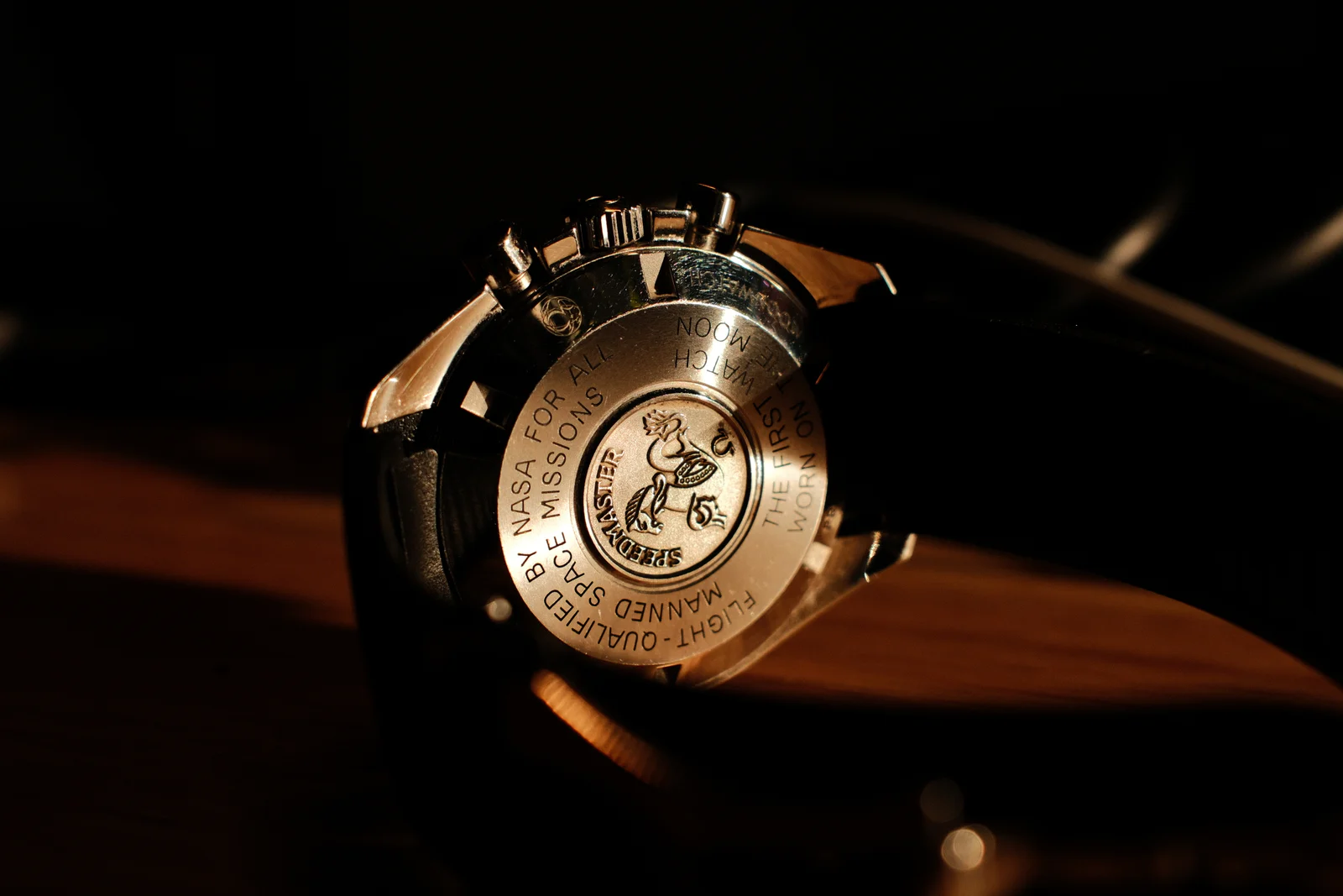 Back of an Omega Speedmaster wristwatch, its steel caseback engraved with a seahorse emblem and NASA flight-qualification text, warmly lit on a wooden surface against darkness.