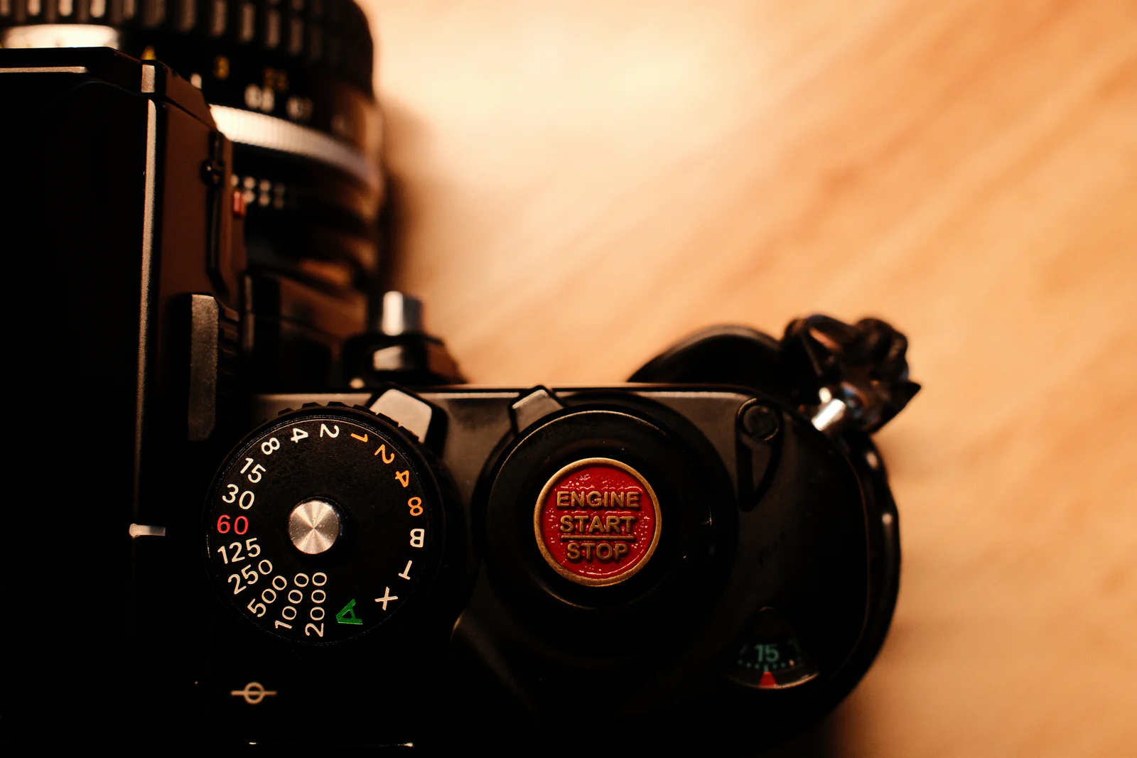 Close-up of a black vintage film camera top showing a shutter speed dial and a red button reading Engine Start Stop, on a blurred wooden surface.